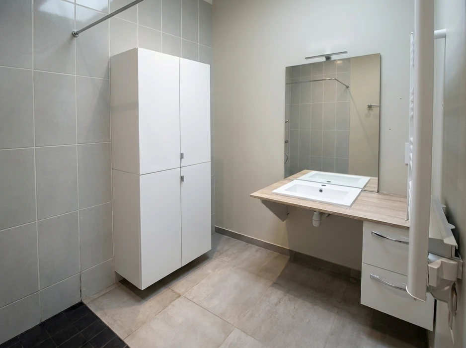 Salle de bain accessible PMR, carrelage gris grand format, vasque encastrée sur plan de travail stratifié bois clair suspendu, miroir avec éclairage LED intégré, armoire de toilette blanche 2 portes, douche à l'italienne avec receveur extra-plat, barre d'appui chromée, tiroirs à galets chromés