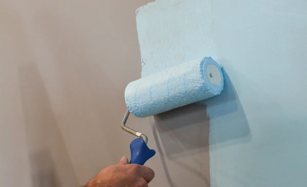 Artisan en salopette et t-shirt blanc installe du papier peint sur mur intérieur résidentiel, appliquant la bande avec les mains près de l'angle et plinthes