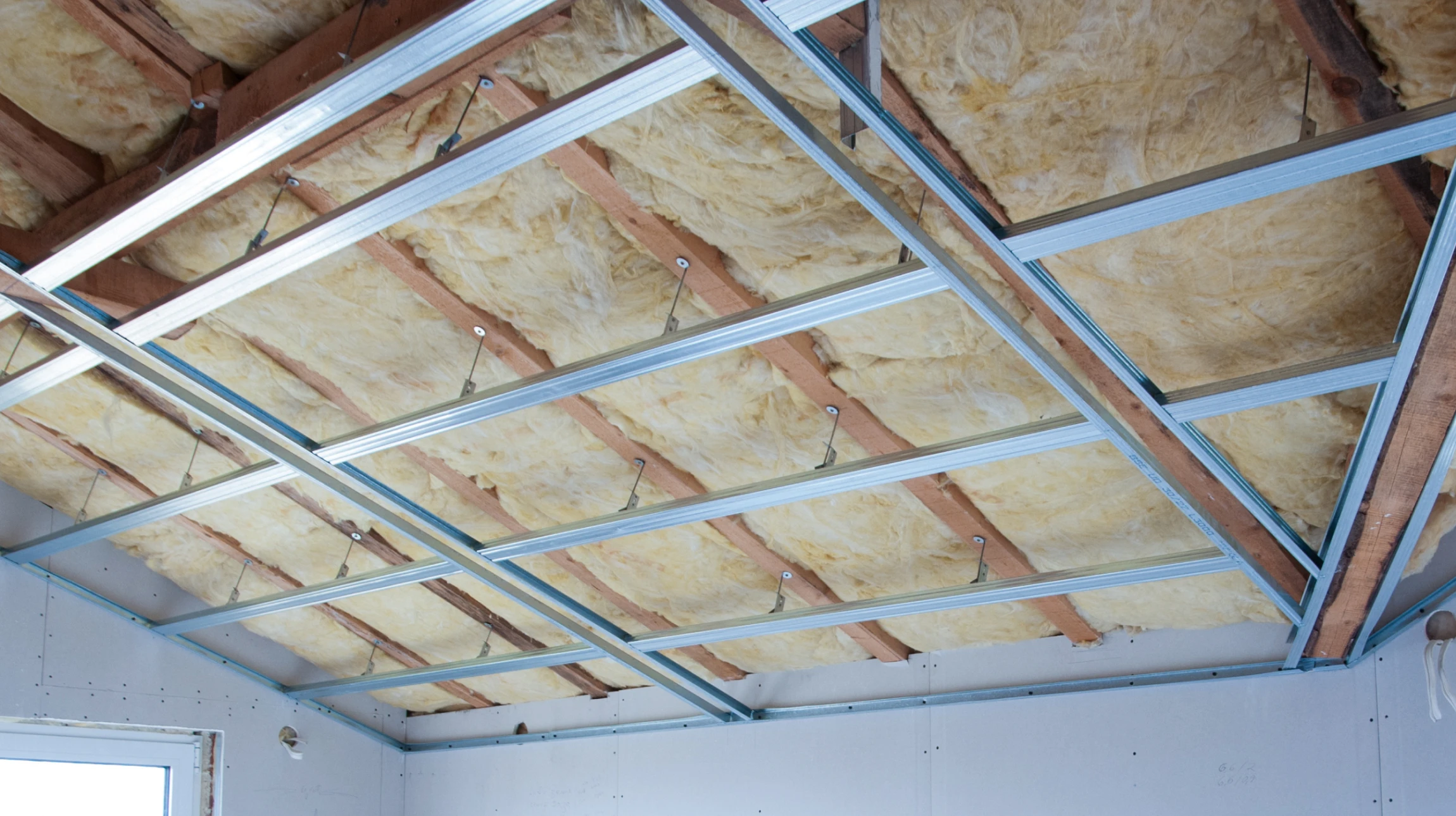 Ouvrier en combinaison bleue installant de l'isolant thermique jaune sous combles, debout sur échelle pliante aluminium
